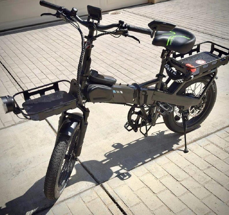 May include: A black electric bicycle with a front basket, rear cargo rack, and a black seat with green accents. The bike has a headlight, fenders, and a kickstand. The tires have a thick tread.