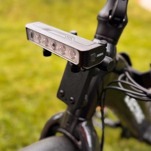 May include: Close-up of a black bicycle headlight with six bright LED lights. The light is mounted on the handlebars of a black bicycle. The background is blurred green grass.