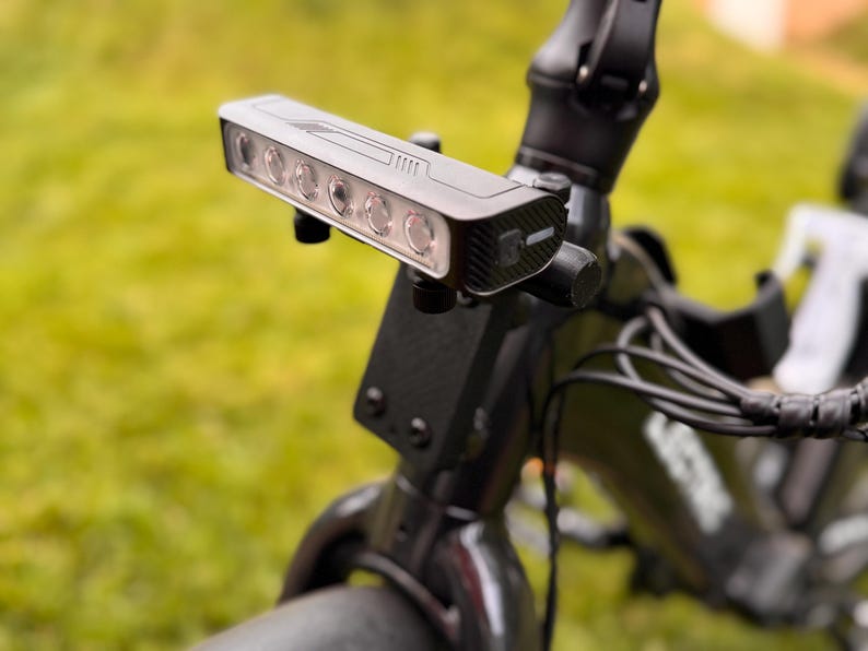 May include: Close-up of a black bicycle headlight with six bright LED lights. The light is mounted on the handlebars of a black bicycle. The background is blurred green grass.