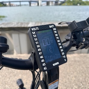 May include: A black electric bike handlebar with a digital display showing the speed, battery level, and voltage. The display reads "00.0" for speed, "00043" for battery level, and "53" for voltage. The display also shows "BATT" for battery and "VOLTS" for voltage.