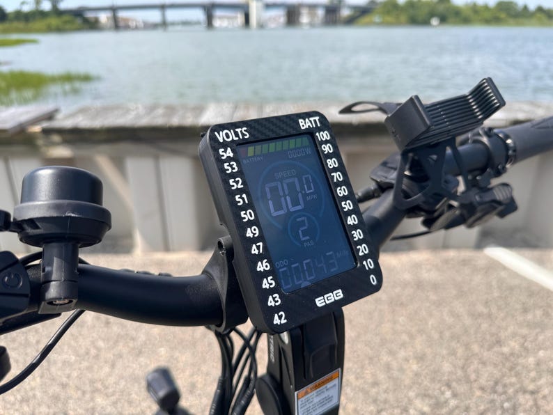 May include: A black electric bike handlebar with a digital display showing the speed, battery level, and voltage. The display reads "00.0" for speed, "00043" for battery level, and "53" for voltage. The display also shows "BATT" for battery and "VOLTS" for voltage.