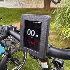 May include: Close-up of an electric bicycle's handlebar with a digital display. The screen shows "00.0 MPH" and "TURBO" in red text. The battery indicator shows 90% charge. The display is mounted on a black textured frame.