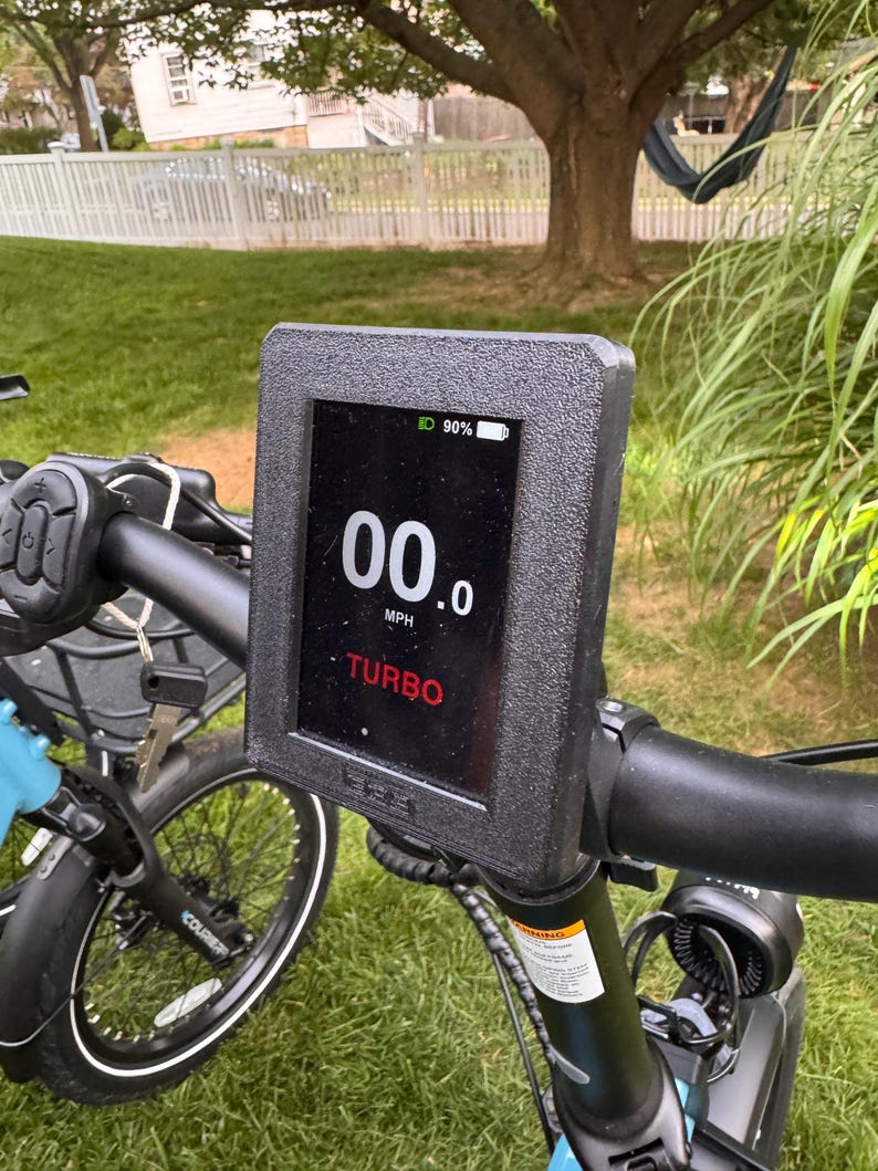 May include: Close-up of an electric bicycle's handlebar with a digital display. The screen shows "00.0 MPH" and "TURBO" in red text. The battery indicator shows 90% charge. The display is mounted on a black textured frame.