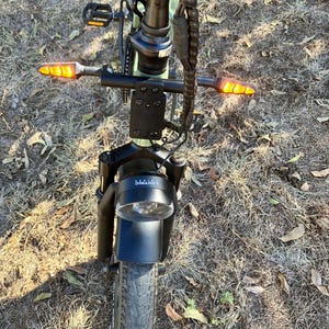 May include: An electric bicycle with a black frame, a round headlight, and amber turn signals. The front tire is thick and black, and the handlebars are black. The bike is on a grassy surface.