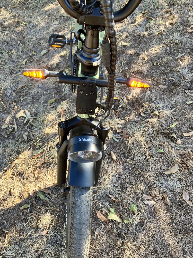 May include: An electric bicycle with a black frame, a round headlight, and amber turn signals. The front tire is thick and black, and the handlebars are black. The bike is on a grassy surface.