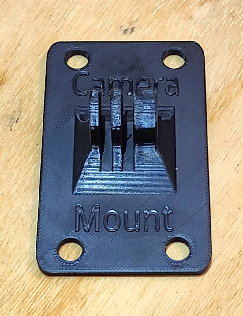 May include: A black 3D printed camera mount with four holes and the text "Camera Mount" printed on it.