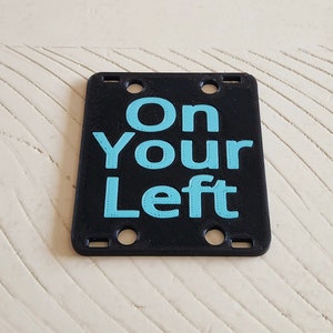 May include: Black plastic sign with four holes. The sign says "On Your Left" in light blue lettering.