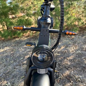 May include: A black and green electric bicycle with a round headlight and orange turn signals. The bicycle has a black handlebar and a front fender. The tires are black and the background is a natural outdoor setting.