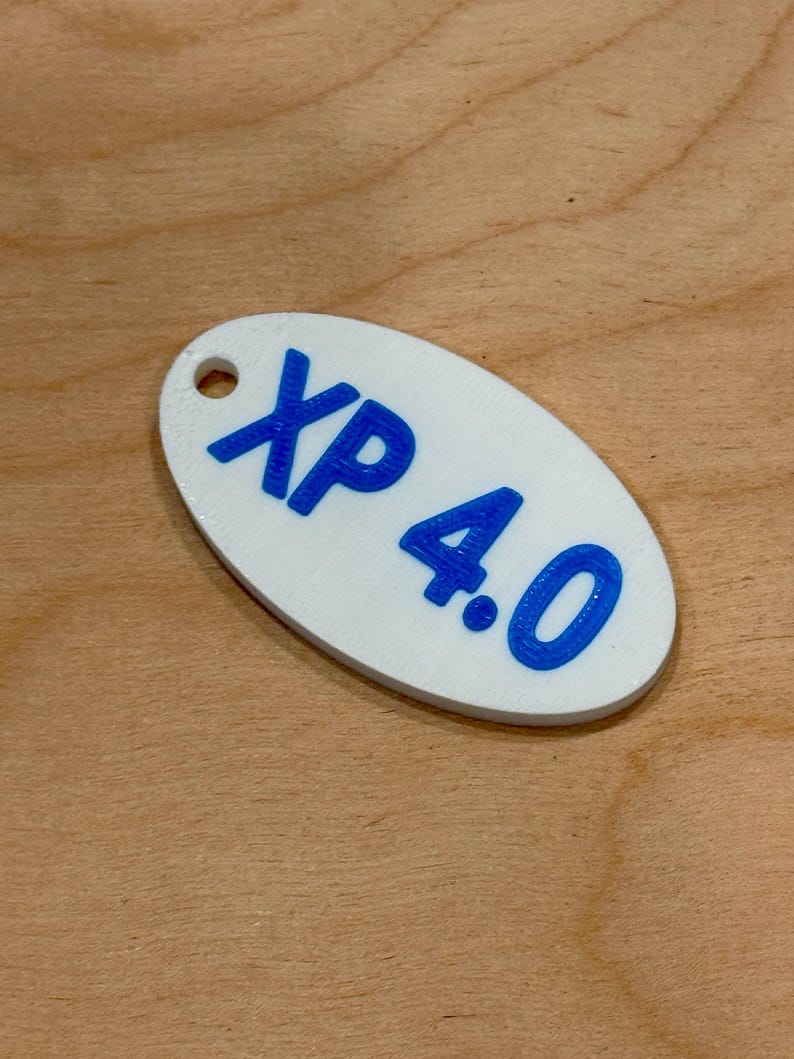 May include: White oval tag with the text "XP 4.0" in blue. The tag has a small hole at the top. The tag is on a wooden surface.