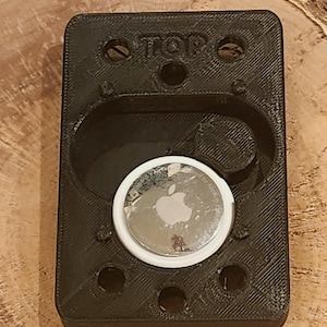 May include: Black rectangular holder with a circular indentation, holding a silver Apple AirTag. The holder has the word "TOP" printed on it and several small holes. The AirTag has the Apple logo.
