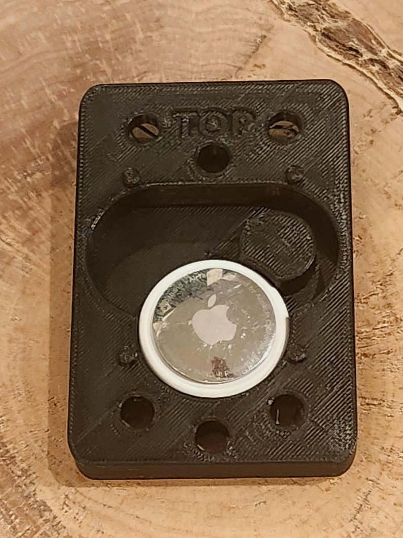 May include: Black rectangular holder with a circular indentation, holding a silver Apple AirTag. The holder has the word "TOP" printed on it and several small holes. The AirTag has the Apple logo.