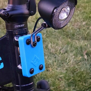 May include: A black bicycle headlight with a blue plastic mount attached to the handlebars. The headlight has a black plastic cover and a clear lens. The mount has a logo on it that says "EG".