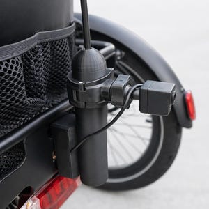 May include: A black bicycle with a rear-mounted camera and a small American flag. The camera is attached to a black pole and is connected by a black wire. The bicycle has a black fender and a red taillight.