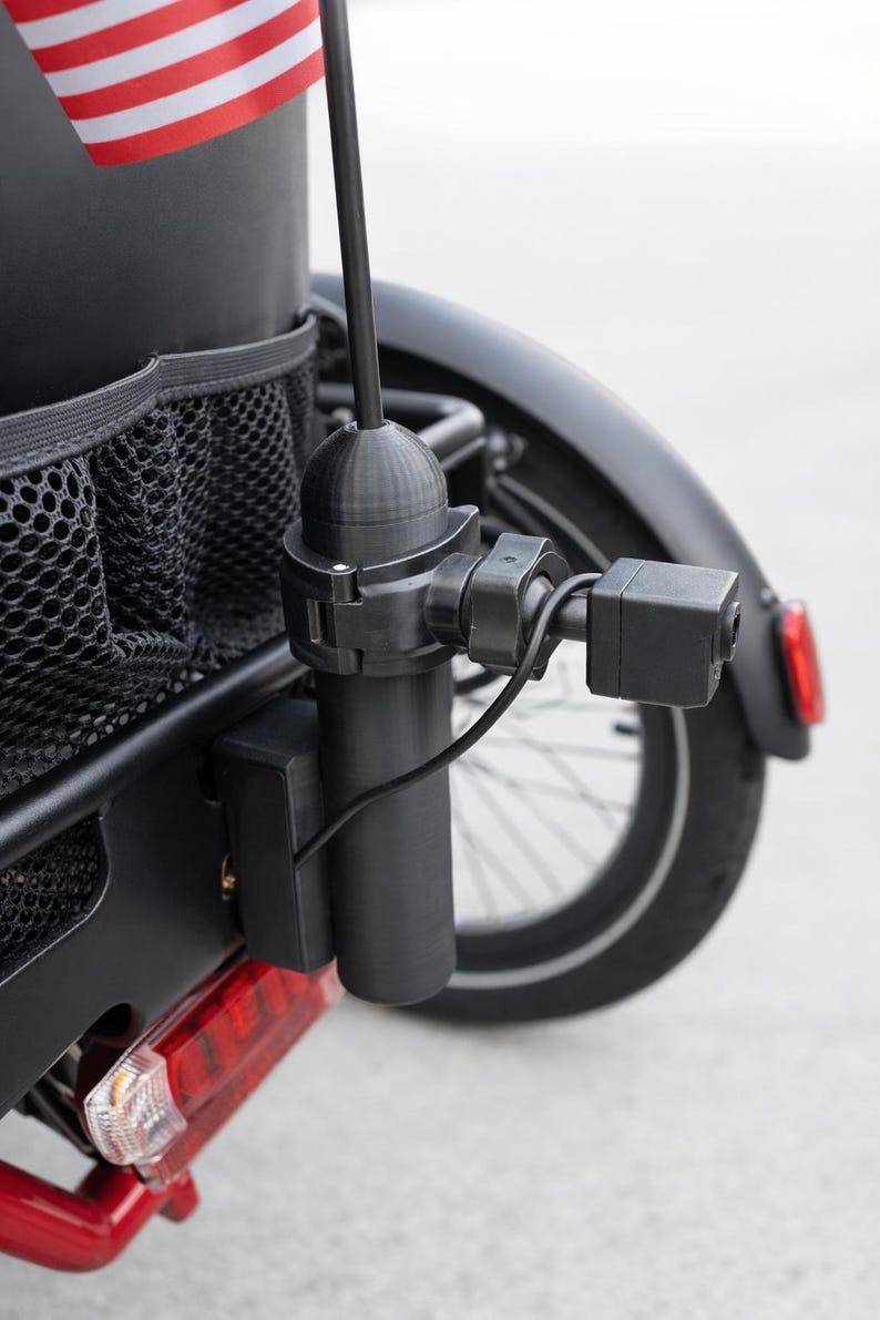 May include: A black bicycle with a rear-mounted camera and a small American flag. The camera is attached to a black pole and is connected by a black wire. The bicycle has a black fender and a red taillight.
