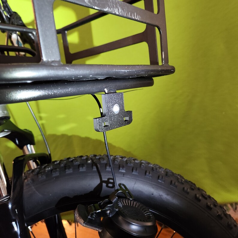 May include: A black metal bike basket with a black plastic mounting bracket attached to the rear of a bicycle. The basket is empty.