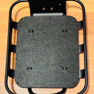 May include: A black metal bicycle rack with a textured grey platform. The rack has a rectangular shape with rounded corners and a curved handle. The platform has several small holes for mounting. The rack is designed to be mounted on a bicycle.