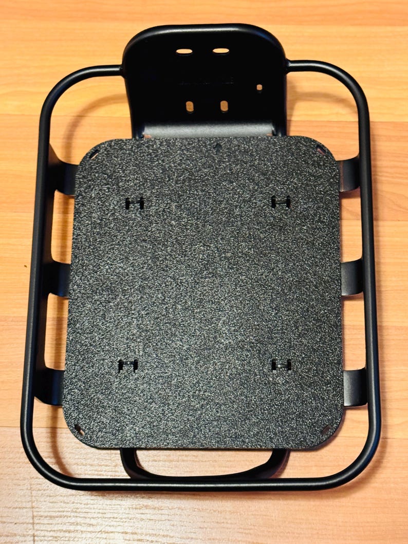 May include: A black metal bicycle rack with a textured grey platform. The rack has a rectangular shape with rounded corners and a curved handle. The platform has several small holes for mounting. The rack is designed to be mounted on a bicycle.