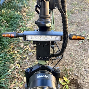 May include: Close-up of an electric scooter's handlebars, featuring a black frame with a headlight and a row of white lights. Amber turn signals extend from the sides. The brand name "i-lectric" is visible on the front.