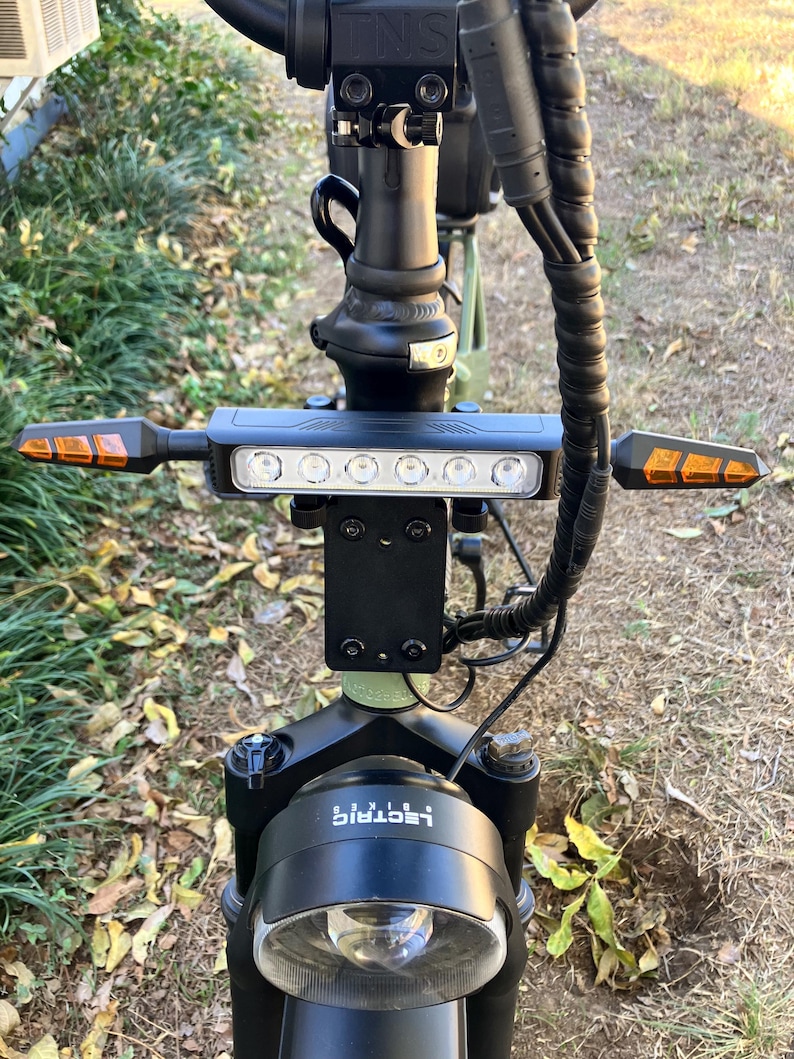 May include: Close-up of an electric scooter's handlebars, featuring a black frame with a headlight and a row of white lights. Amber turn signals extend from the sides. The brand name "i-lectric" is visible on the front.
