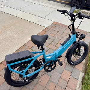 May include: A teal Lectric XP 2.0 electric bike with black tires, seat, and battery pack. The bike has a digital display, front headlight, and a black handlebar. The bike is parked on a brick and concrete surface.
