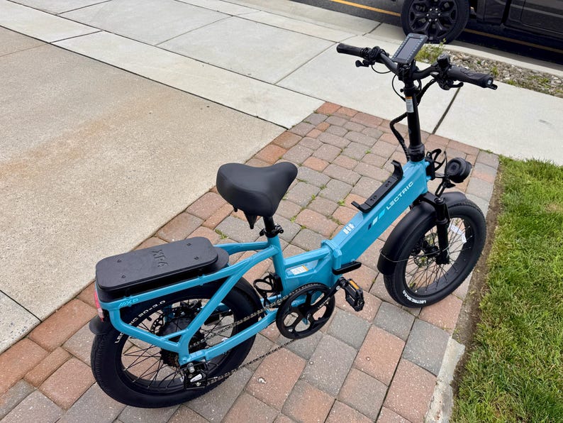 May include: A teal Lectric XP 2.0 electric bike with black tires, seat, and battery pack. The bike has a digital display, front headlight, and a black handlebar. The bike is parked on a brick and concrete surface.
