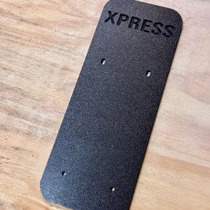 May include: A black rectangular sign with the word "XPRESS" embossed in white letters. The sign has four small holes drilled into it.