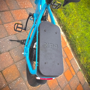 May include: A turquoise electric bicycle with a black seat and pedals. A black rectangular panel with the text "XP4" is mounted on the rear. The bike is parked on a brick surface with green grass in the background.