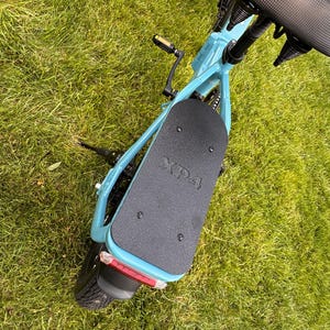 May include: A light blue electric scooter with a black seat and footboard. The footboard has the text "XP4" printed on it. The scooter is on a grassy surface.