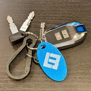 May include: A collection of keys and a car key fob on a dark surface. The keys include two metal keys and a key with a black plastic head. A blue oval keychain with a white logo is also present. The car key fob is black and silver.