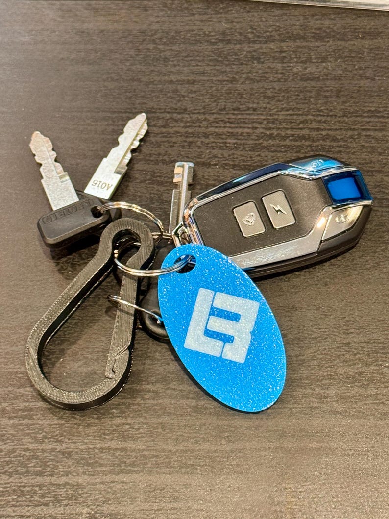 May include: A collection of keys and a car key fob on a dark surface. The keys include two metal keys and a key with a black plastic head. A blue oval keychain with a white logo is also present. The car key fob is black and silver.