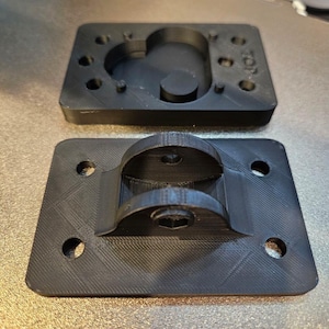May include: Two black, rectangular plastic components. The top piece has a curved recess and several small holes. The bottom piece features a curved bracket and mounting holes. Both pieces have a textured surface.