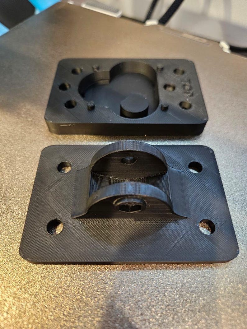 May include: Two black, rectangular plastic components. The top piece has a curved recess and several small holes. The bottom piece features a curved bracket and mounting holes. Both pieces have a textured surface.