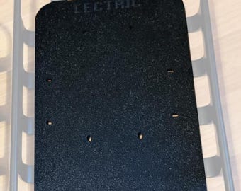 May include: A black rectangular plate with the word "LECTRIC" printed on it, placed inside a gray bicycle basket. The plate has a textured surface and several small holes.