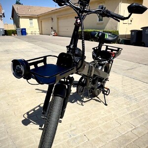 May include: Black electric bicycle with a front basket, headlight, and rear cargo rack. The bike has a black seat with green accents, black handlebars, and a digital display. The tires are thick and black, suitable for various terrains.