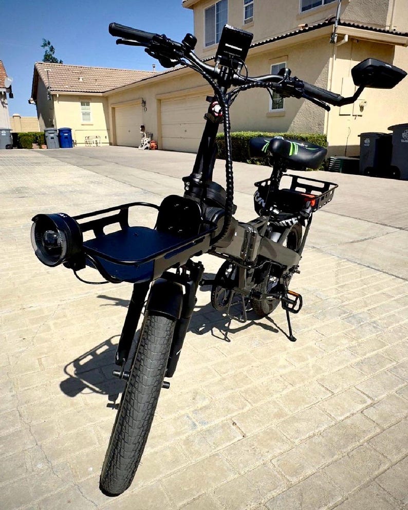 May include: Black electric bicycle with a front basket, headlight, and rear cargo rack. The bike has a black seat with green accents, black handlebars, and a digital display. The tires are thick and black, suitable for various terrains.