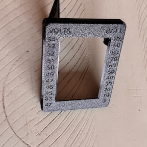 May include: A black, rectangular voltmeter housing with the words "VOLTS" and "BATT" printed on it. The housing has a series of numbers indicating voltage and battery percentage. The device is made of a textured material and is mounted on a wall.