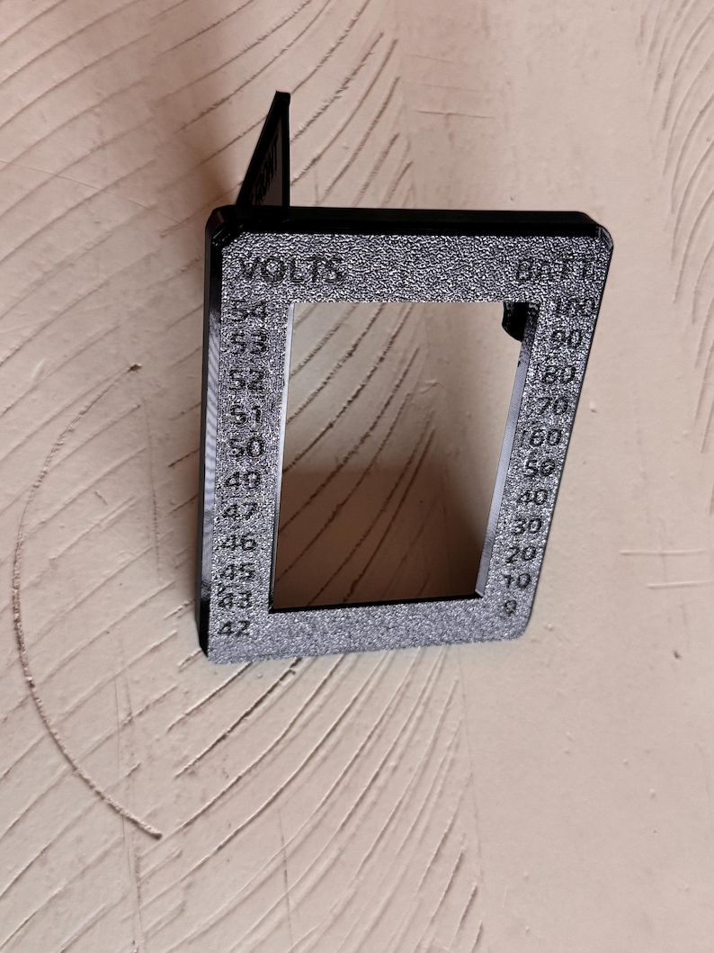 May include: A black, rectangular voltmeter housing with the words "VOLTS" and "BATT" printed on it. The housing has a series of numbers indicating voltage and battery percentage. The device is made of a textured material and is mounted on a wall.
