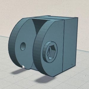 May include: A 3D-rendered, teal-colored mechanical component. The design features a rectangular base with two circular, semi-circular elements on one side. One circular element has a hole, and the other has a hexagonal socket. A triangular cutout is visible.