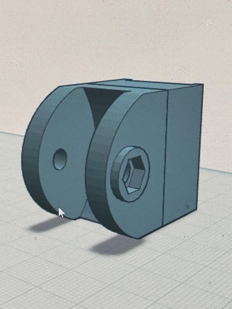 May include: A 3D-rendered, teal-colored mechanical component. The design features a rectangular base with two circular, semi-circular elements on one side. One circular element has a hole, and the other has a hexagonal socket. A triangular cutout is visible.