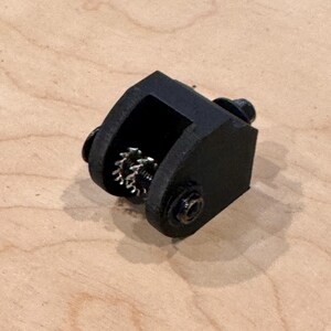 May include: A black, 3D-printed mechanical component with a gear visible inside a rectangular housing. The component is secured with small black screws. The object is resting on a light brown wooden surface.