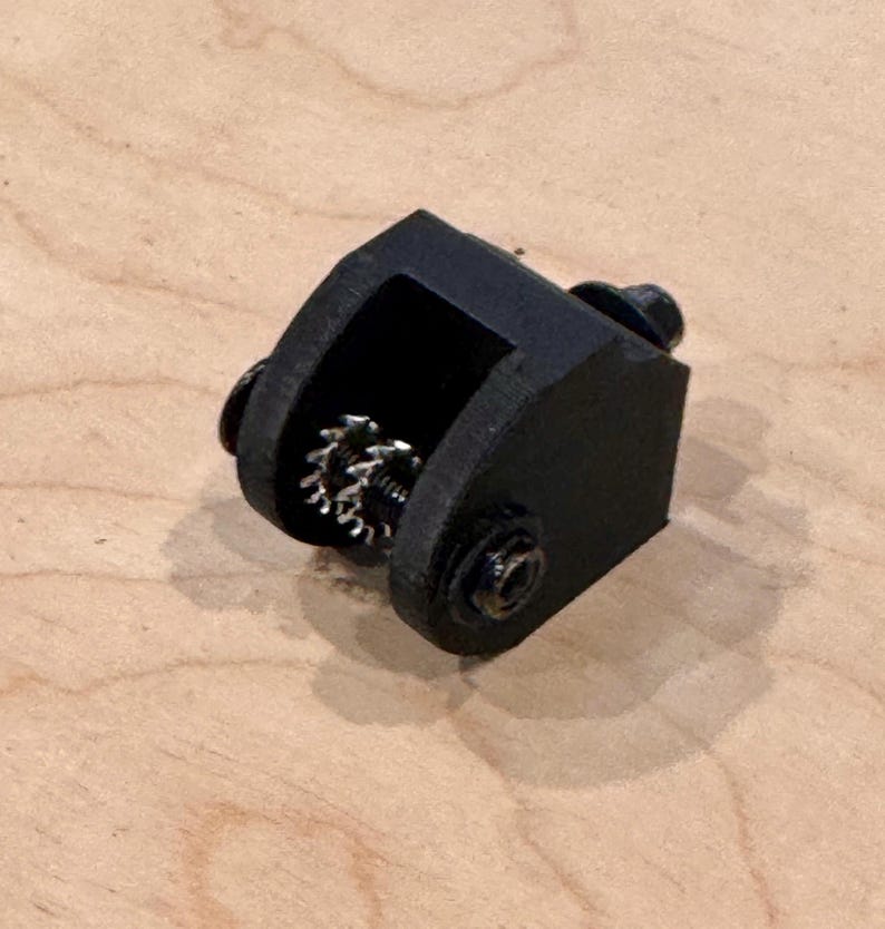 May include: A black, 3D-printed mechanical component with a gear visible inside a rectangular housing. The component is secured with small black screws. The object is resting on a light brown wooden surface.