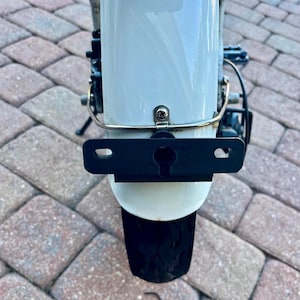 May include: A white bicycle fender with a black license plate holder attached to it. The fender is mounted on a bicycle with a black tire.