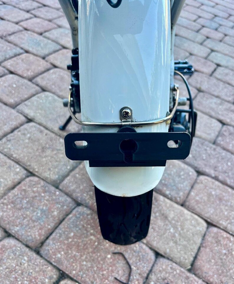 May include: A white bicycle fender with a black license plate holder attached to it. The fender is mounted on a bicycle with a black tire.