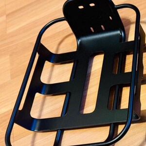 May include: A black metal bicycle basket with a backrest. The basket has a rectangular shape with cut-out sections and a curved frame. The backrest has three small holes. The basket is designed to be mounted on a bicycle.