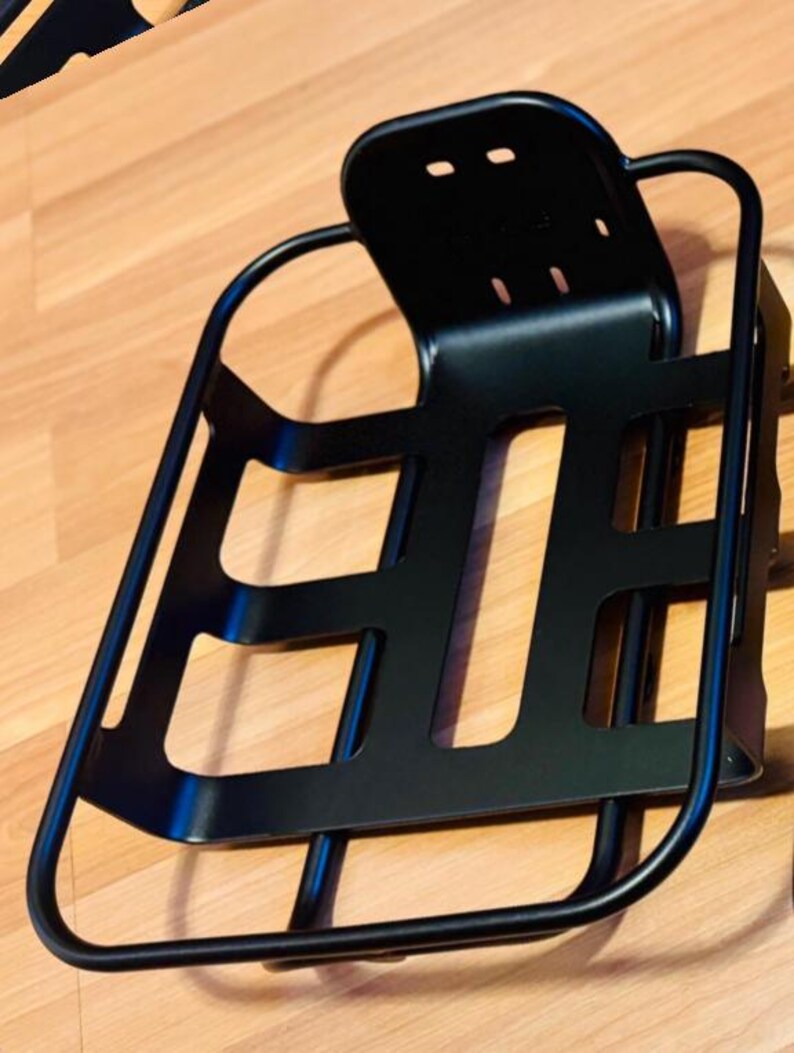 May include: A black metal bicycle basket with a backrest. The basket has a rectangular shape with cut-out sections and a curved frame. The backrest has three small holes. The basket is designed to be mounted on a bicycle.