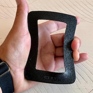 May include: A black, rectangular, textured object with rounded corners is held in a hand. The object has a cutout center and appears to be made of a matte material. The hand is holding the object against a light-colored surface.