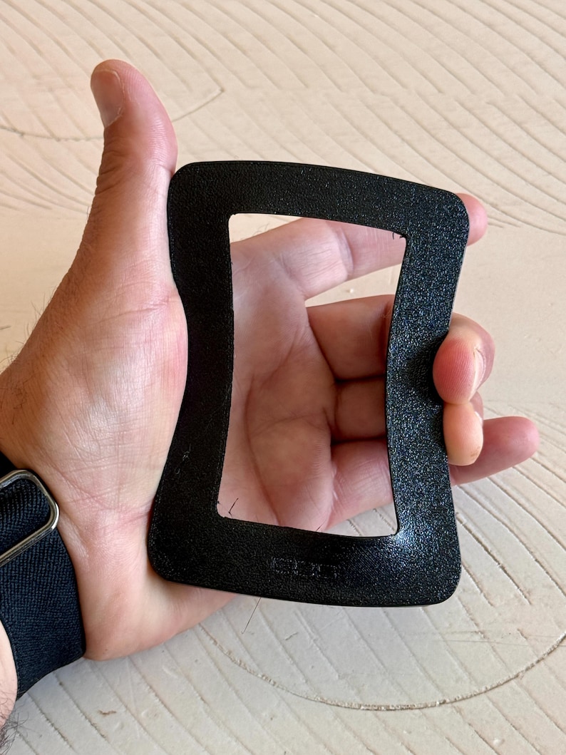 May include: A black, rectangular, textured object with rounded corners is held in a hand. The object has a cutout center and appears to be made of a matte material. The hand is holding the object against a light-colored surface.