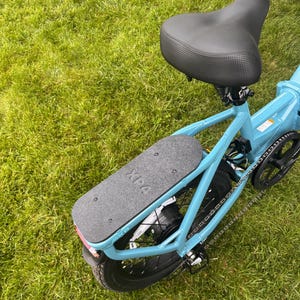 May include: A light blue electric bicycle with a black seat and a black footboard. The footboard has the text "XPA" printed on it. The bicycle is on a green grassy surface.