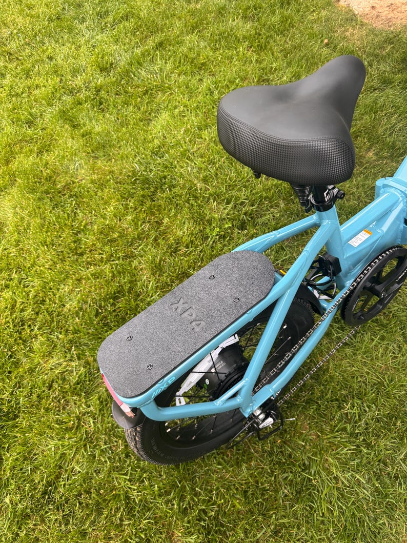 May include: A light blue electric bicycle with a black seat and a black footboard. The footboard has the text "XPA" printed on it. The bicycle is on a green grassy surface.