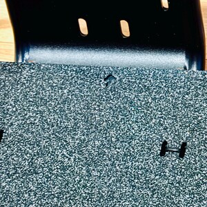 May include: Close-up of a textured, speckled, grey surface with a black metal component. The metal piece has two oval cutouts and a curved shape. The surface has a small, diamond-shaped cutout and a black, H-shaped piece.